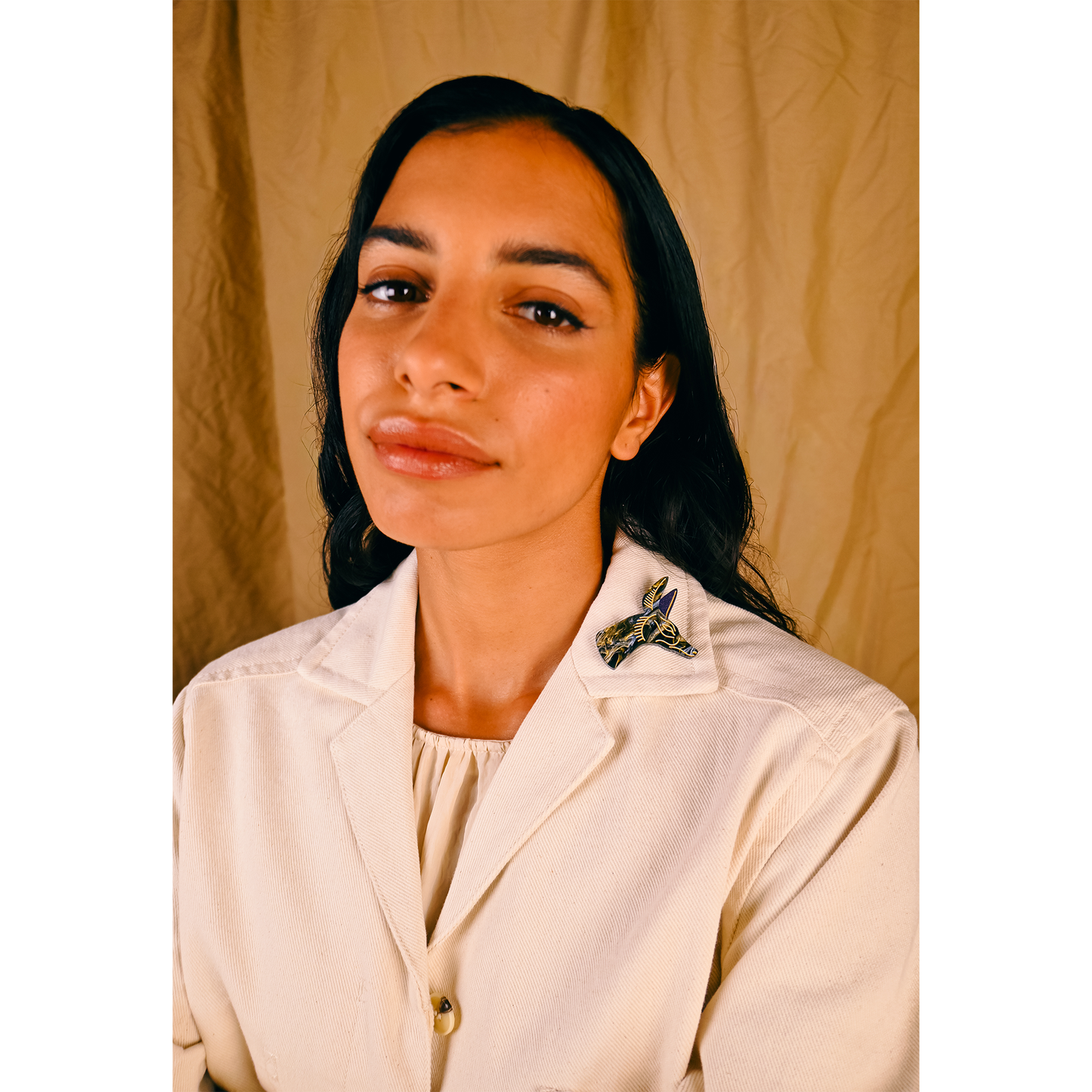 Woman wearing a beige shirt with a decorative gold and black Egyptian jackal brooch against a beige background