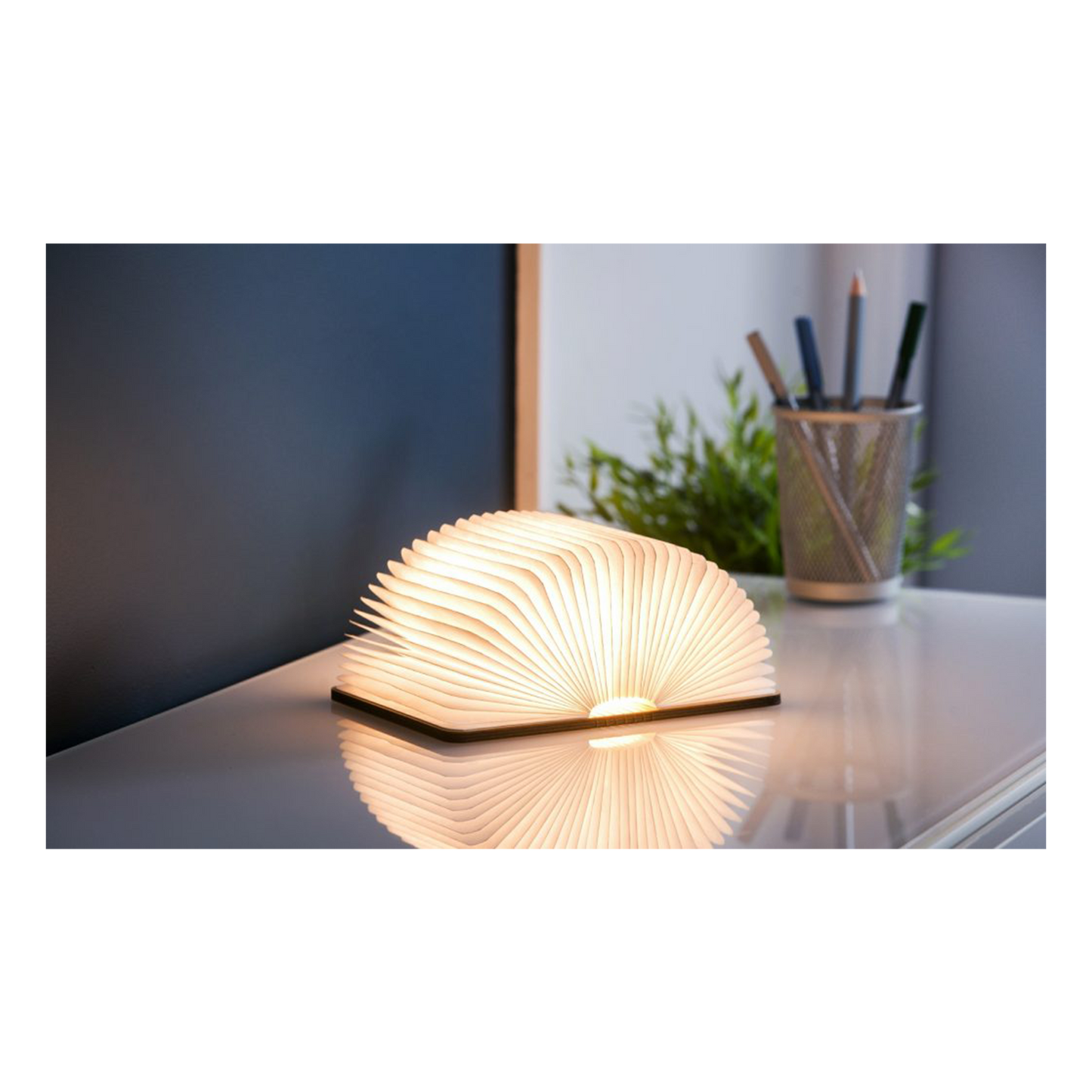 Book-shaped lamp on a desk with a plant and pen holder in the background