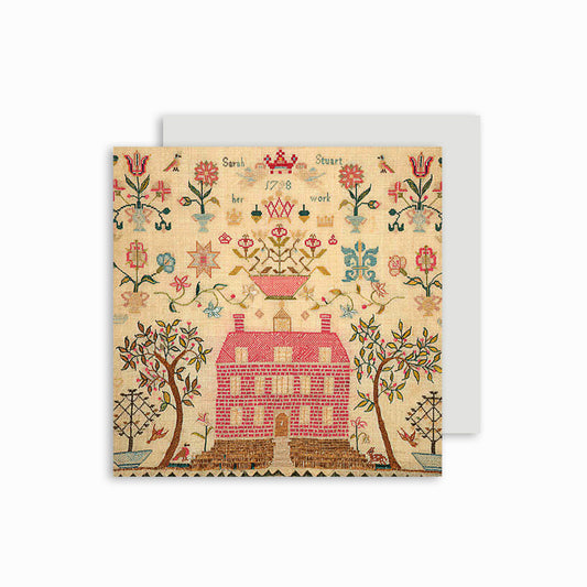 A greeting card featuring a historical needlework sampler design with a red house, surrounded by floral patterns. The card is cello wrapped with a white envelope displayed behind.